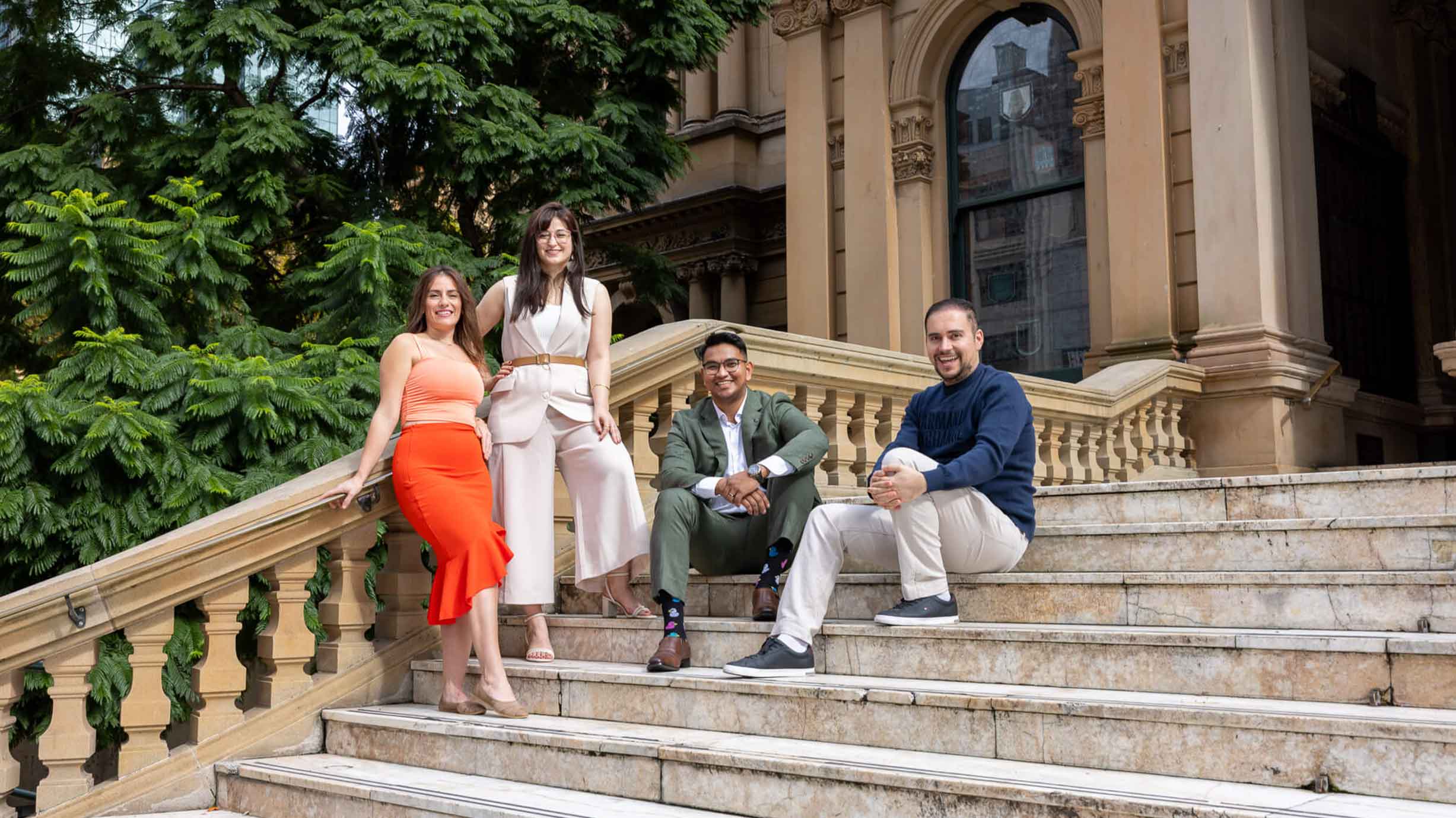 Four adults pose together on outdoor marble steps in front of a historic building with arched windows and lush greenery.